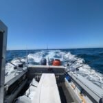 Fishing charter boat cruising offshore with multiple trolling rods set and ocean wake behind.