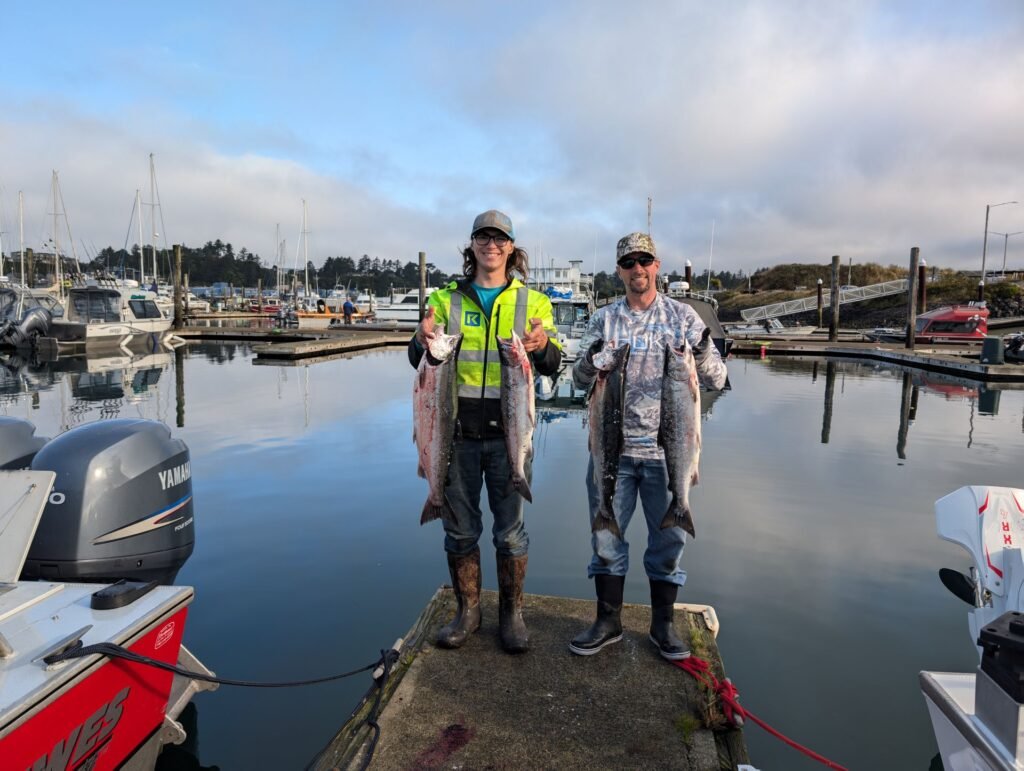 Two anglers standing on a marina dock in Newport Oregon holding freshly caught salmon from a fishing trip.