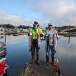 Two anglers standing on a marina dock in Newport Oregon holding freshly caught salmon from a fishing trip.